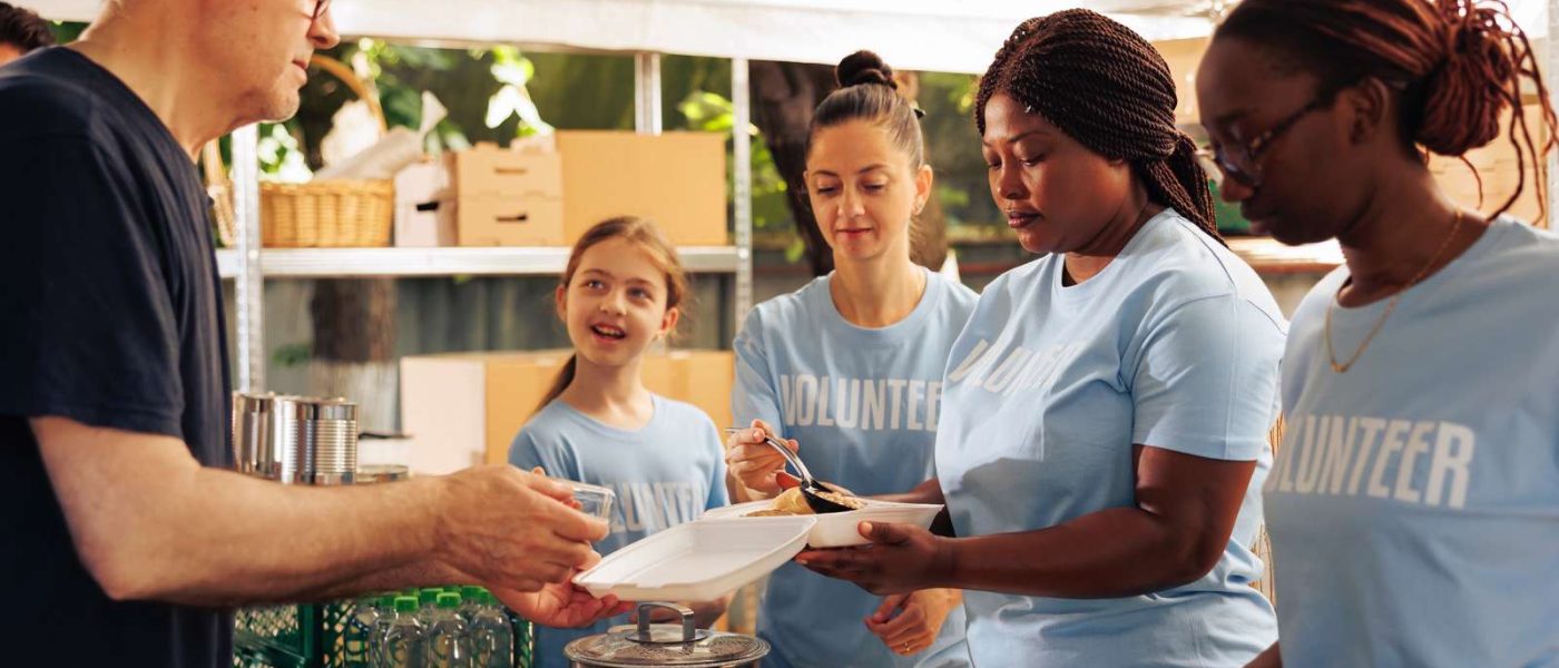 Multiracial volunteer women at local center giving food assistance serving meals and non-perishable items to hungry and homeless people. Friendly charity workers feed warm meals to underprivileged.